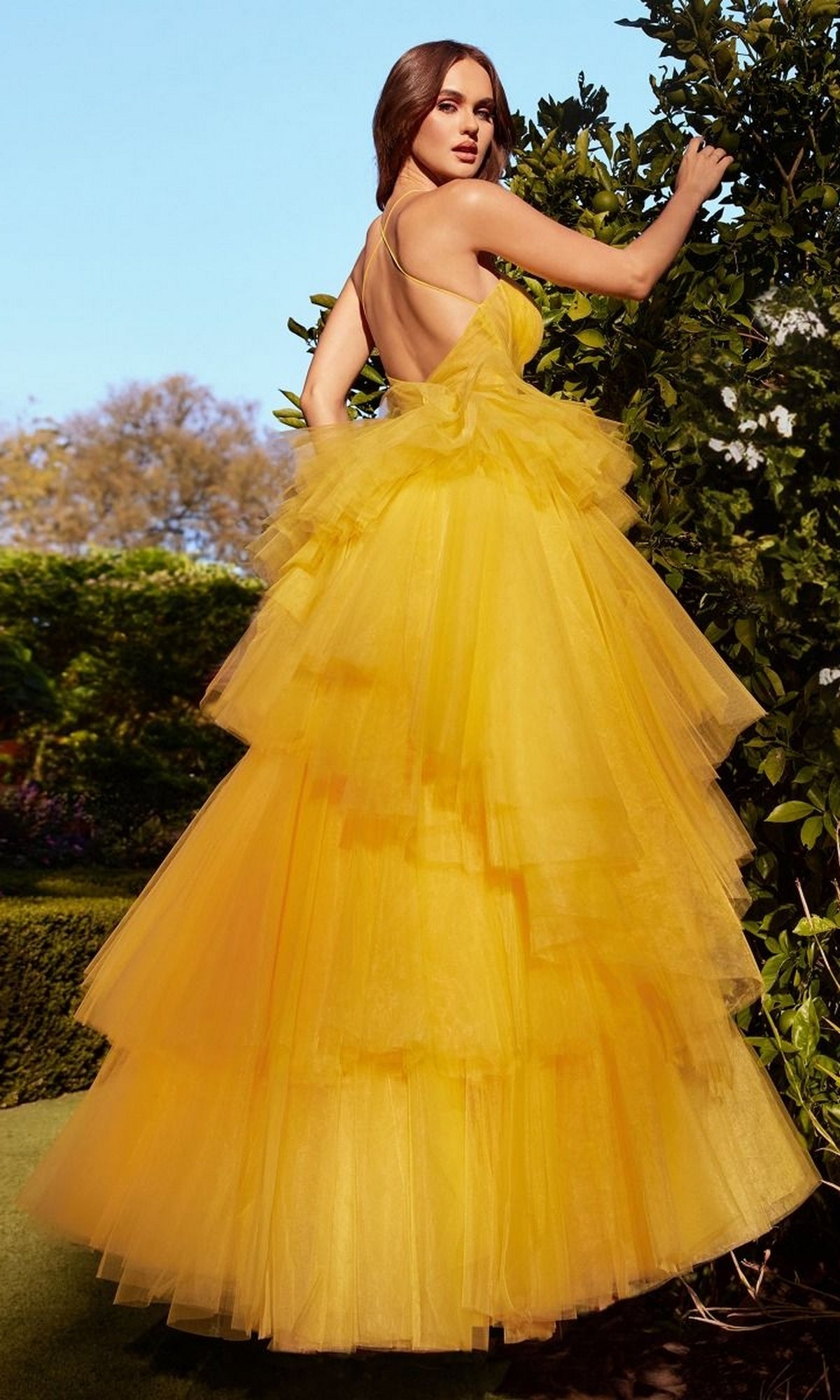 A woman stands outdoors among green foliage and trees, wearing the Bright Cut-Out Long Tiered Prom Ball Gown A1238—a vibrant yellow dress with an open back and tiered design.